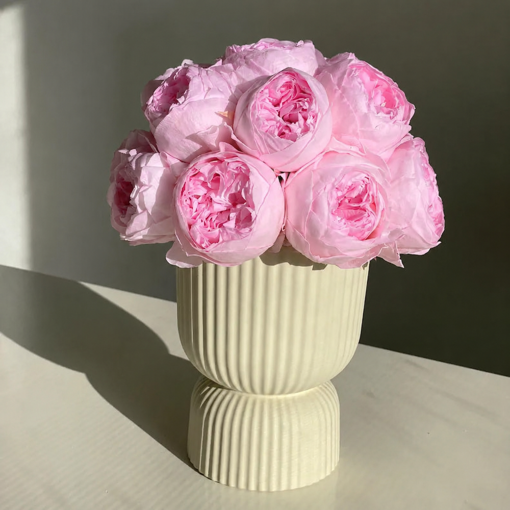 A bouquet of light pink peony flowers arranged in a ribbed cream ceramic vase, set on a light surface against a neutral background.