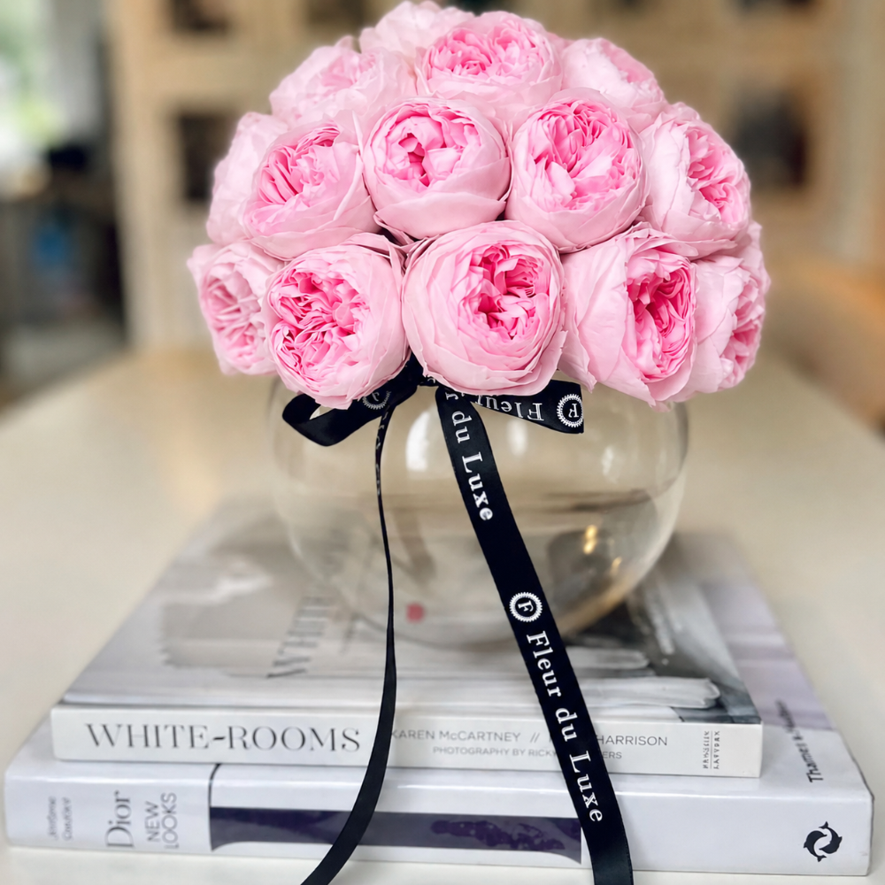 A bouquet of pink peony-like flowers arranged in a clear round glass vase, tied with a black ribbon printed with white text, resting on stacked white books.