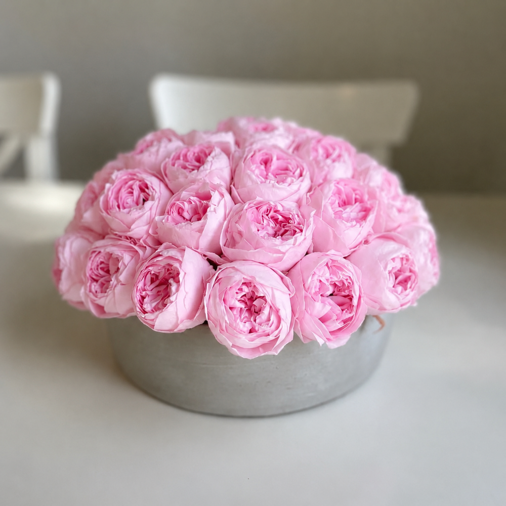 A round arrangement of light pink peony roses in a gray round pot, set on a light-colored table with a softly blurred indoor background.