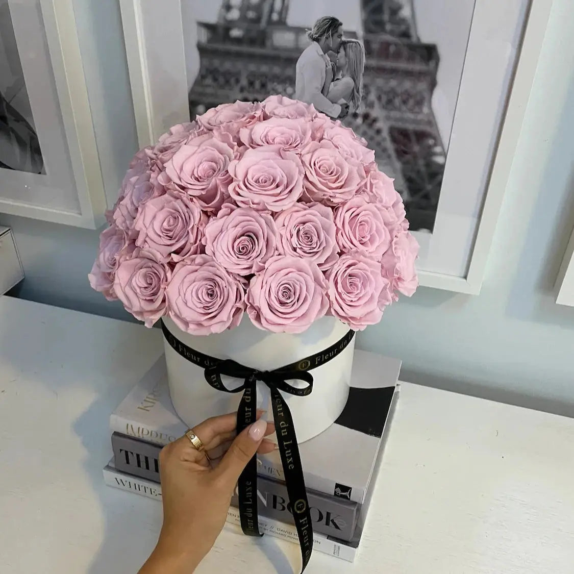 Pink roses in a white box with a black ribbon, held by a hand against a light blue wall with framed pictures.