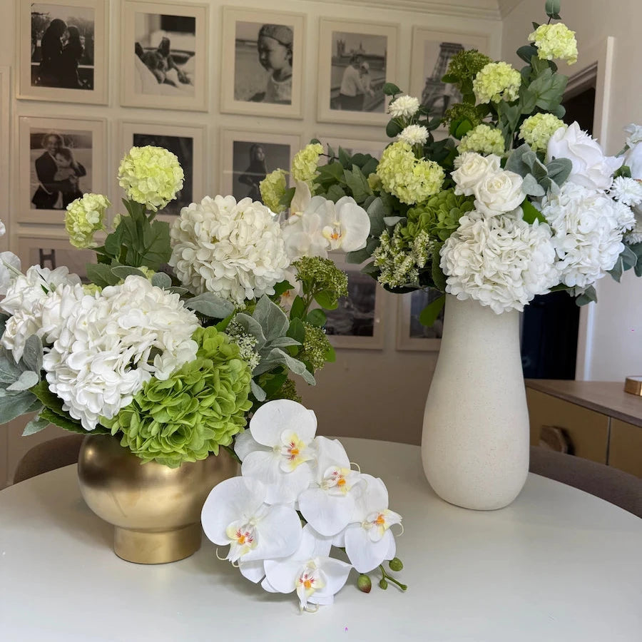 Decorative arrangement of flowers in vases on a table with a wall of framed photos in the background.