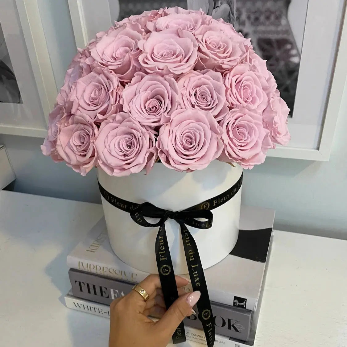 Bouquet of pink roses in a white box with a black ribbon, held by a hand on top of books.
