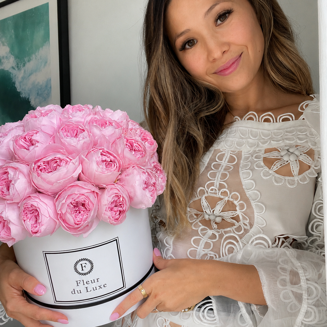 A woman in a white lace dress holds a round white box filled with pink roses. The box has a label that reads 'Fleur du Luxe'.
