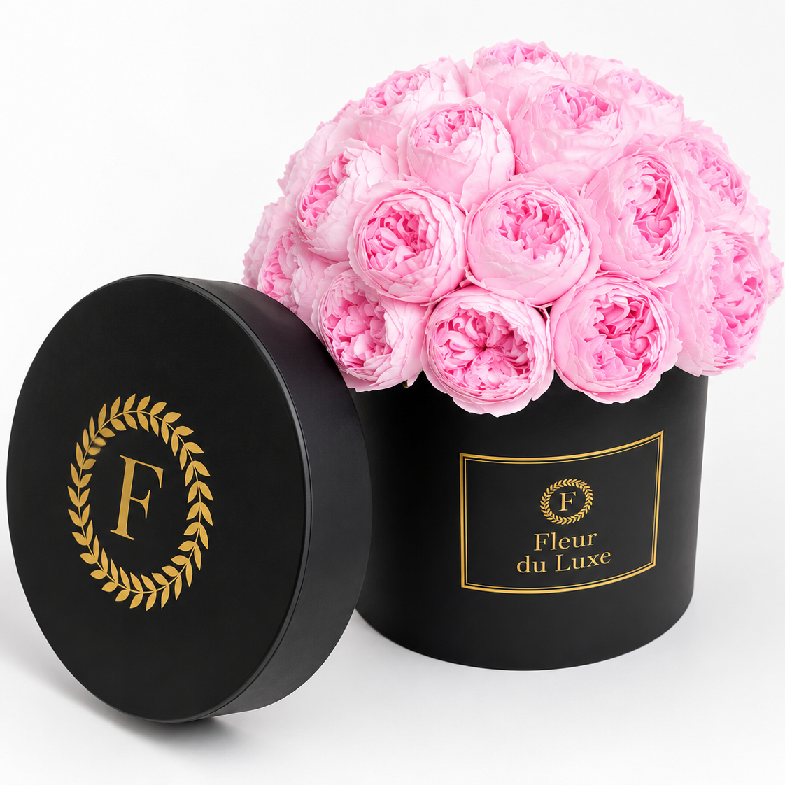A round black flower box with its lid leaning beside it, filled with a bouquet of pink peony flowers. The box is branded with a gold monogram 'F' wreath and the text 'Fleur du Luxe'.