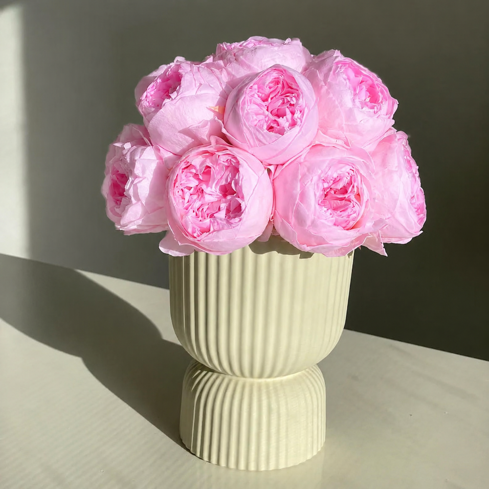 A bouquet of pink peony-like artificial flowers arranged in a ribbed cream-colored ceramic vase, set on a light surface against a neutral background.