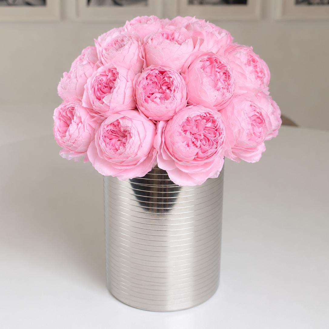 A bouquet of light pink peony flowers arranged in a ribbed silver cylindrical vase, set on a white tabletop with a softly blurred indoor background.