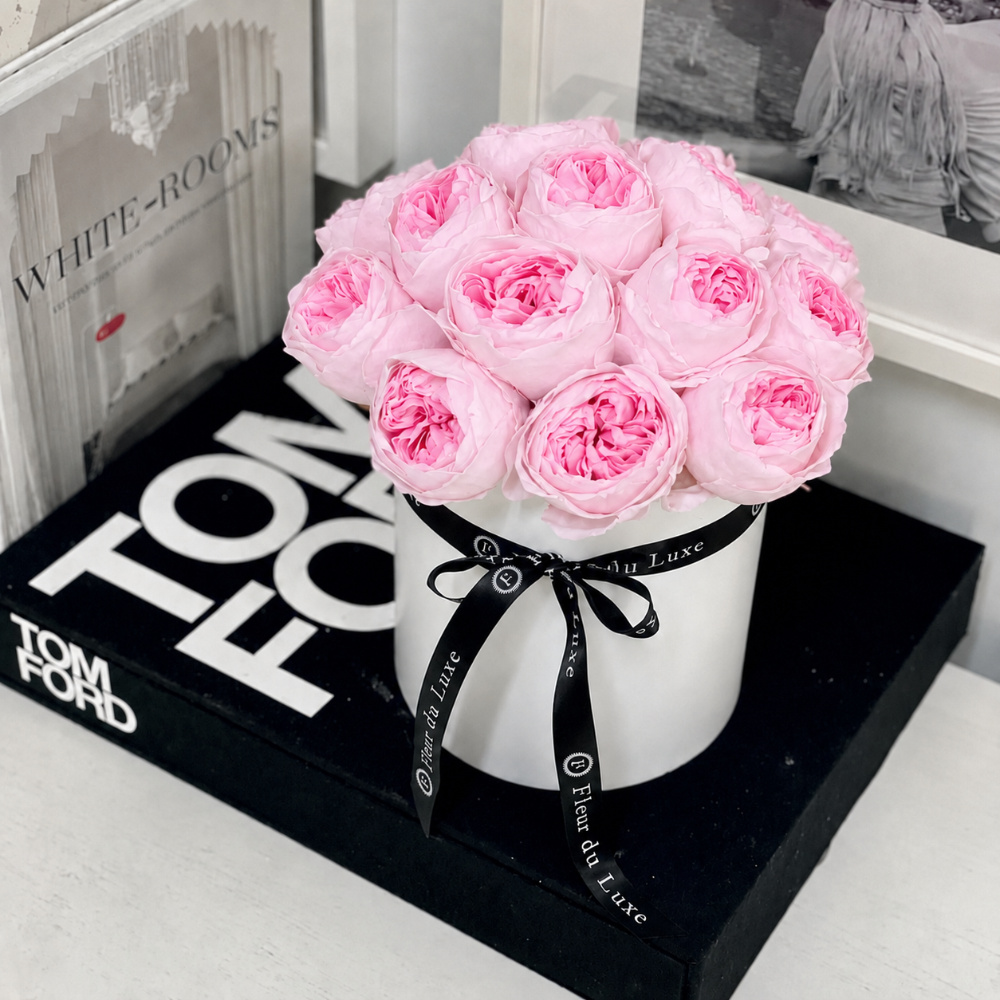 A bouquet of light pink roses arranged in a white cylindrical box tied with a black ribbon printed with 'Tom Ford Luxe', placed on a black Tom Ford box in an indoor setting.
