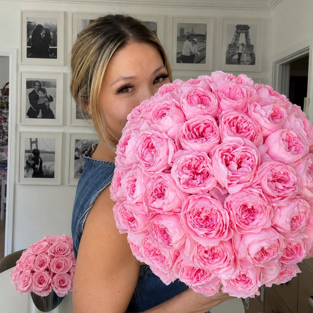 A woman in a denim top holds a large, dense bouquet of light pink roses in front of her face, with a smaller matching bouquet on a table in the background.